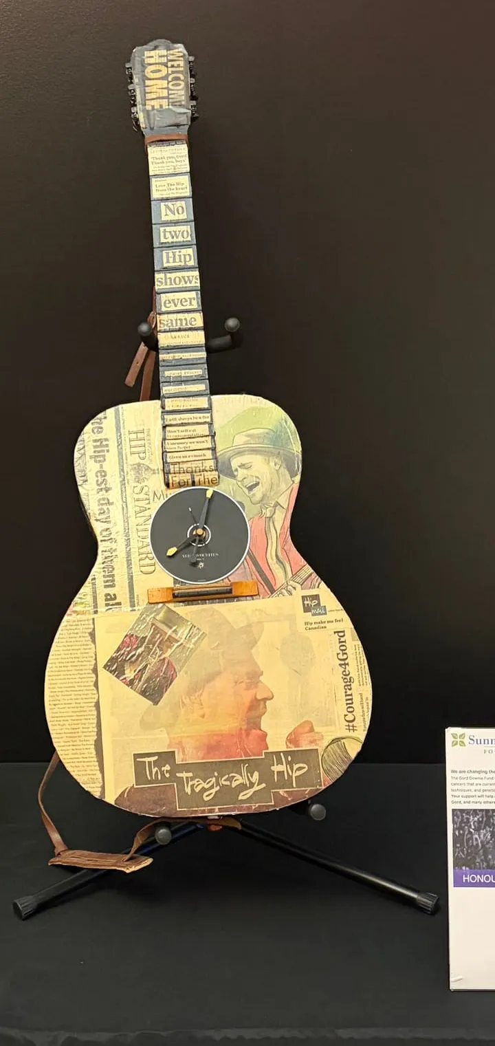 Guitar-shaped tribute clock with portraits, lettering and The Tragically Hip name, displayed at a fundraising event.