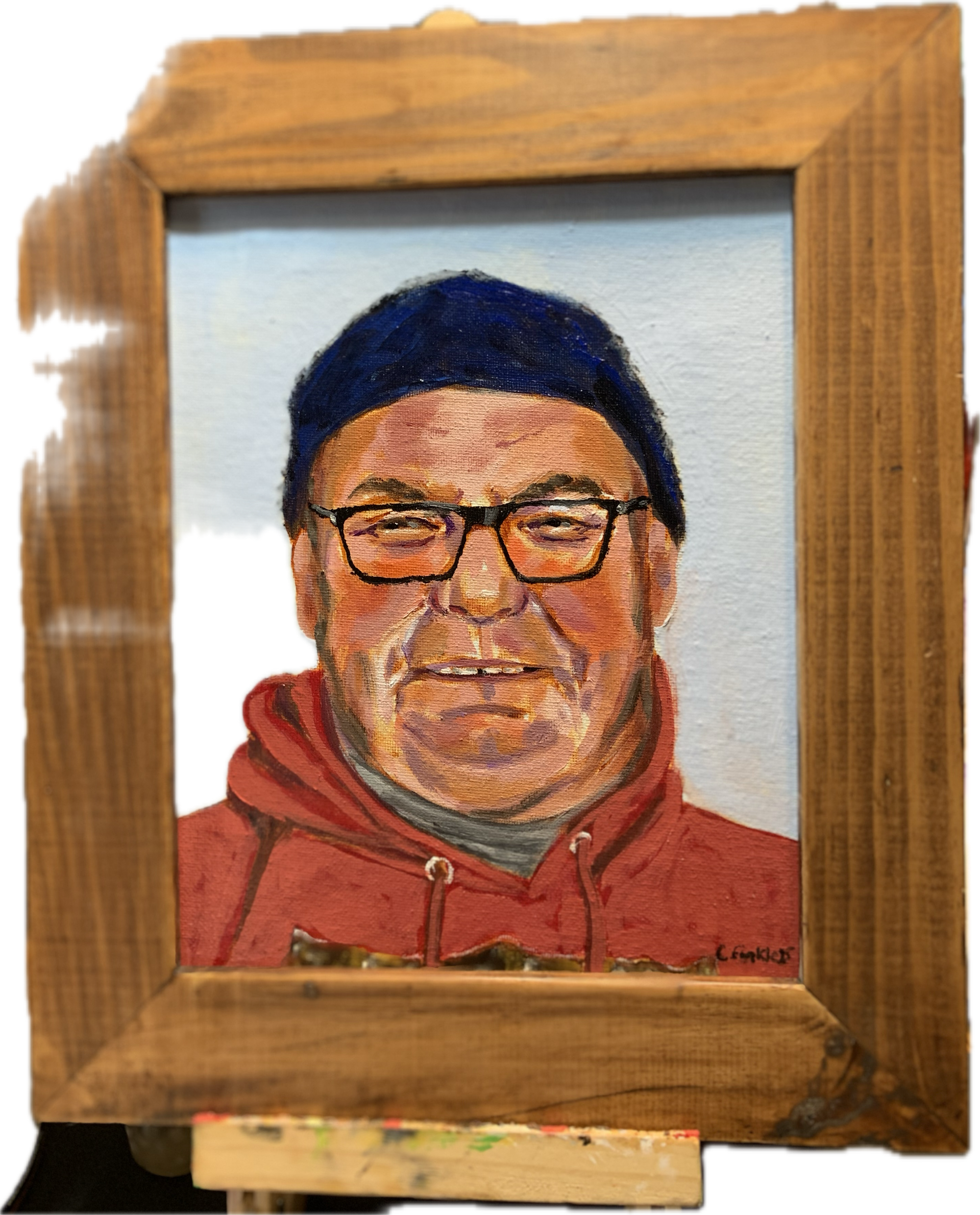 Framed portrait painting of a man in glasses, a dark cap and a red hoodie.