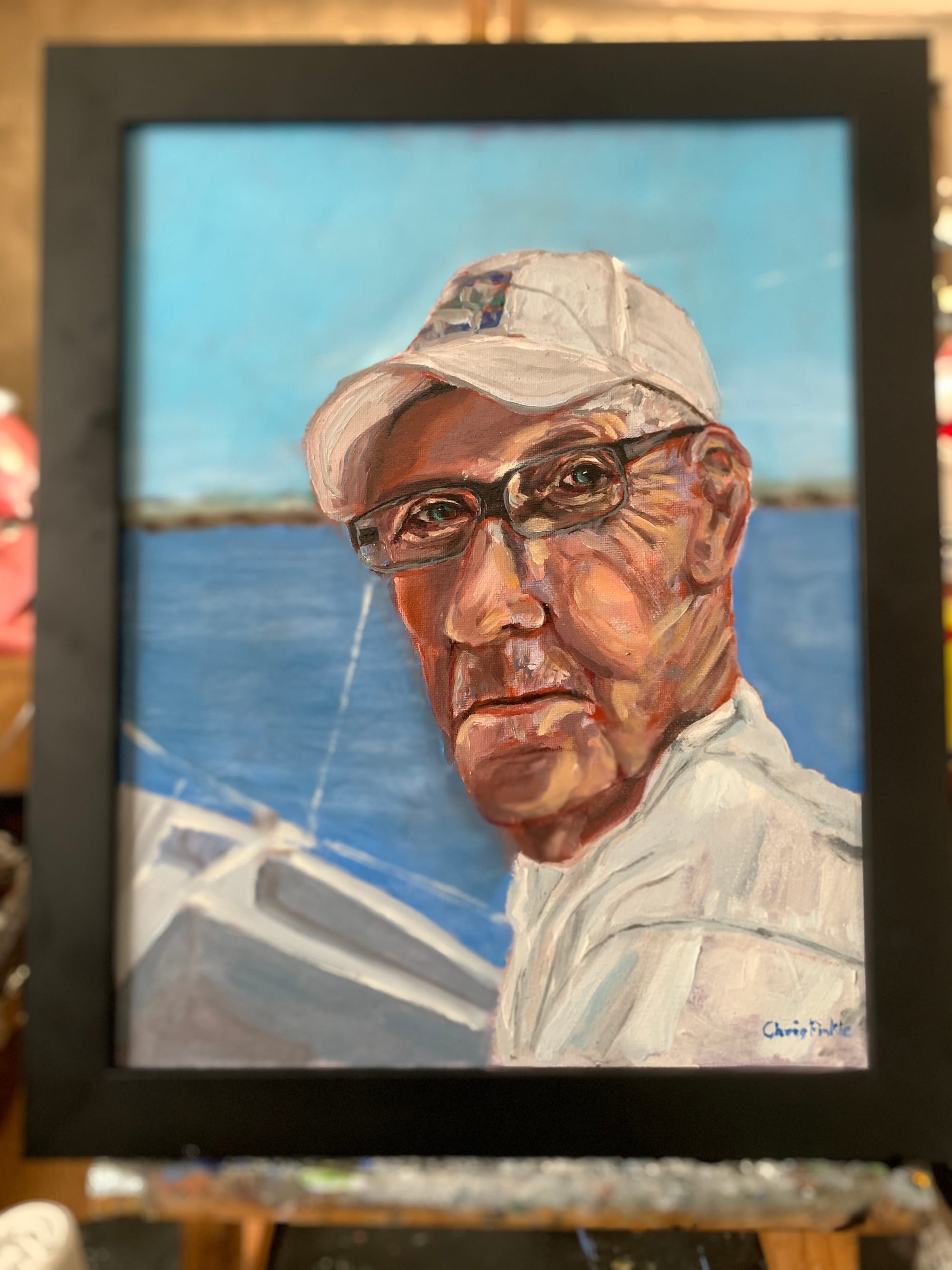 Framed portrait painting of a man wearing a light cap and dark glasses beside the water.