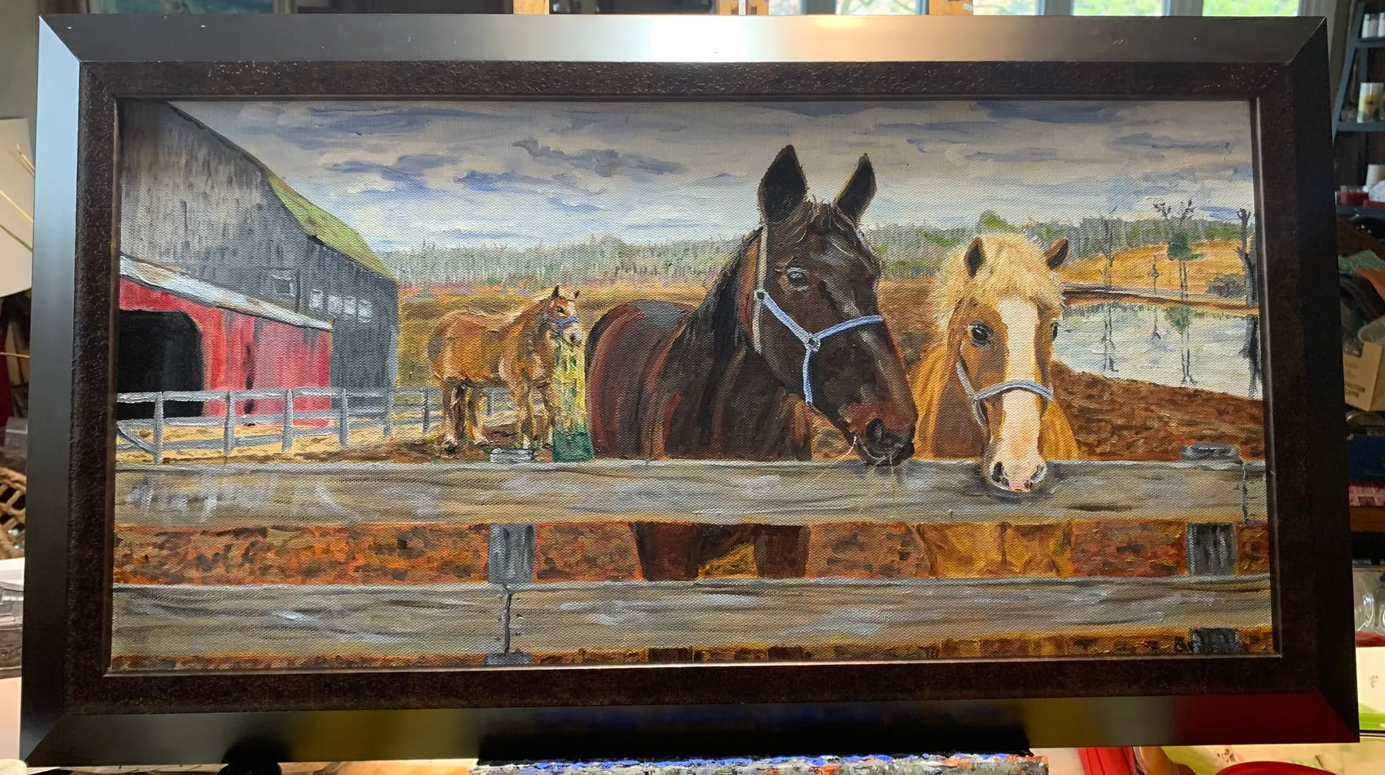 Framed landscape painting of horses standing at a wooden fence beside a barn.