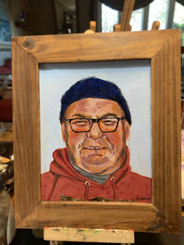 Framed portrait painting of Ernie wearing a red hoodie, dark cap and glasses.